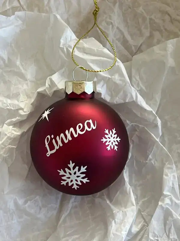 En Personlig julkula i glas från Expresspresenter.se i rött med vita snöflingor och namnet Linnea vilar på skrynklat vitt papper. Upptill sitter en guldfärgad hängtråd på denna unika juldekoration.