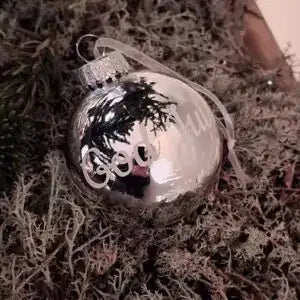Julgranskula i silver med egen text från Expresspresenter.se, personifierad med "Good", vilar på mossa och grönska och dess blanka yta reflekterar omgivande grenar.