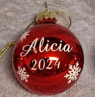 En 6 cm bred rödglänsande "Plastkula till julgran med egen text" från Expresspresenter.se, med "Alicia 2024" i vit skrift och snöflingor, vilar på en grå yta med struktur.