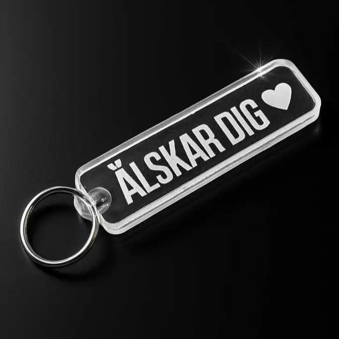 Nyckelringen Älskar dig från Express-presenter är en rektangulär nyckelring i akryl med metallring, där "ÄLSKAR DIG" och ett hjärta graverat i vitt, "love you", visas på en svart yta.