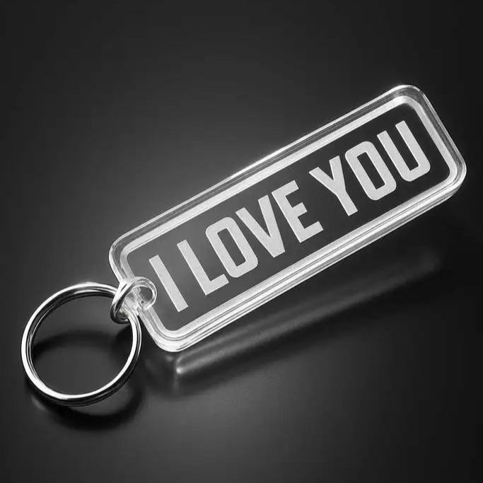 Nyckelringen I love you by Express-presenter är en rektangulär nyckelring i akryl med en metallring, som visar "I LOVE YOU" i feta vita bokstäver på svart bakgrund, visad på en mörk yta.