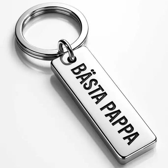 Express-presenter Nyckelring | Bästa pappa är en silverfärgad nyckelring med en rektangulär tagg i rostfritt stål med texten "BÄSTA PAPPA" ingraverad på en vit yta.