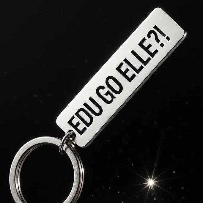 En silverfärgad Nyckelring | EDU GO ELLE?! från Express-presenter vilar på en mörk, glittrande yta och visar upp en rektangulär rostfritt stål tagg med laseretsad text: EDU GO ELLE?!.