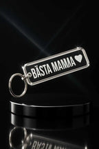 En blank akrylnyckelring från Express-presenter med "Bästa mamma" och en hjärtsymbol, på en svart rund piedestal mot en mörk bakgrund. Den graverade baksidan ger denna Nyckelring Bästa mamma en elegant och omtänksam touch.