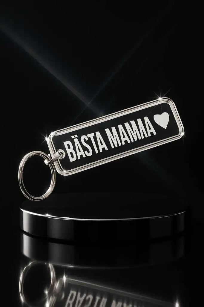 En blank akrylnyckelring från Express-presenter med "Bästa mamma" och en hjärtsymbol, på en svart rund piedestal mot en mörk bakgrund. Den graverade baksidan ger denna Nyckelring Bästa mamma en elegant och omtänksam touch.