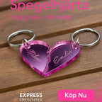 Nyckelring - Halvt hjärta i spegelplast (rosa)" från Express-presenter är en hjärtformad nyckelring (4x3cm) i rosa akryl delad i två med namnen Milda och Emilie ingraverade, visad på trä med svensk text och Köp Nu-knapp.