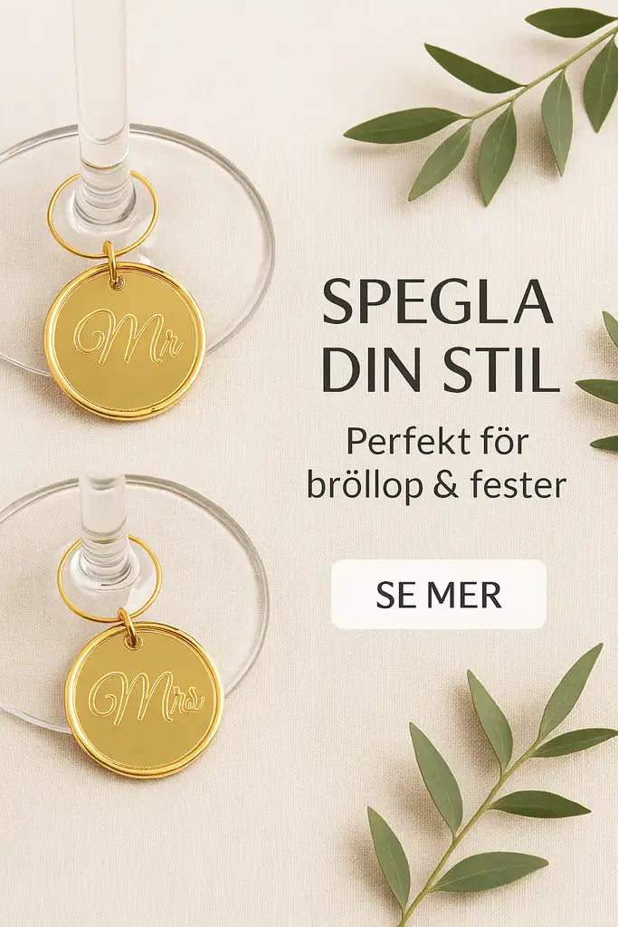 Två vinglas med guld Glasmarkörer bröllop 2st från Expresspresenter.se, märkta Mr och Mrs, vilar på en beige yta med gröna blad. Dessa berlocker i spegelakryl är perfekta för bröllop och fest.