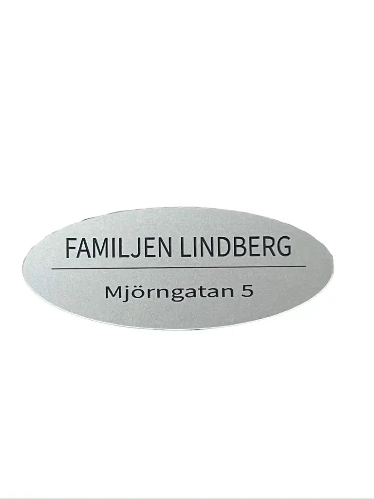 En Stilren brevlådeskylt från Express-presenter, oval i akryl, 110x60 mm, med texten FAMILJEN LINDBERG ovanför en linje och Mjörngatan 5 under.