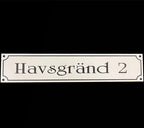 Adresskylt-Gatuvägskylt by Express-presenter är en vit rektangulär skylt (300×100 mm) tillverkad av slitstark akrylmaterial med "Havsgränd 2" i svart text på svart botten, dekorativa hörn och två små monteringshål.