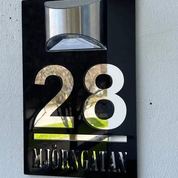 Express-presenter's Husnummerskylt med belysning i svart och silver visar "28 MJÖRNGATAN" på en vit vägg, med en modern armatur ovanför för snygg belysning.