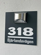 En svart Husskylt med belysning by Express-presenter, med vit text "318" och "Bjärelandavägen", är monterad på en ljusvägg med en silverfärgad utomhusarmatur ovanför som dekorativ belysning.