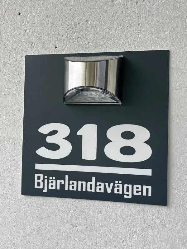 En svart Husskylt med belysning by Express-presenter, med vit text "318" och "Bjärelandavägen", är monterad på en ljusvägg med en silverfärgad utomhusarmatur ovanför som dekorativ belysning.