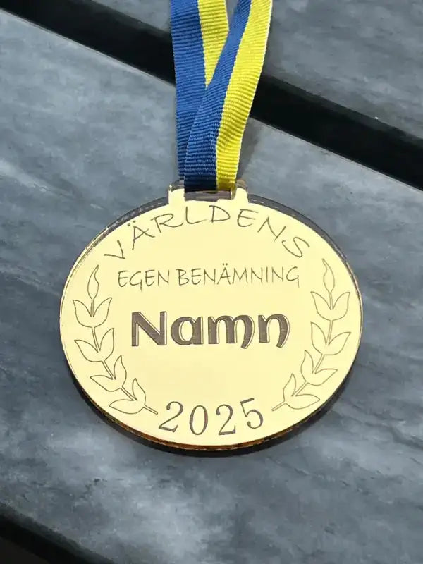 Medaljen Världens egen benämning från Express-presenter, med ett 3mm blågult sverigeband och graverat "Världens egen benämning Namn 2025", vilar på en mörk bänk.
