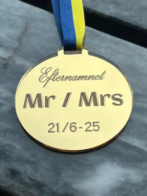 Medalj | Mr / Mrs by Express-presenter är en guldmedalj med ett blågult sverigebandsband, graverad med Efternamnet, Mr / Mrs, och datumet 21/6 - 25. Tillverkad av spegelplast, den vilar på en grå yta.