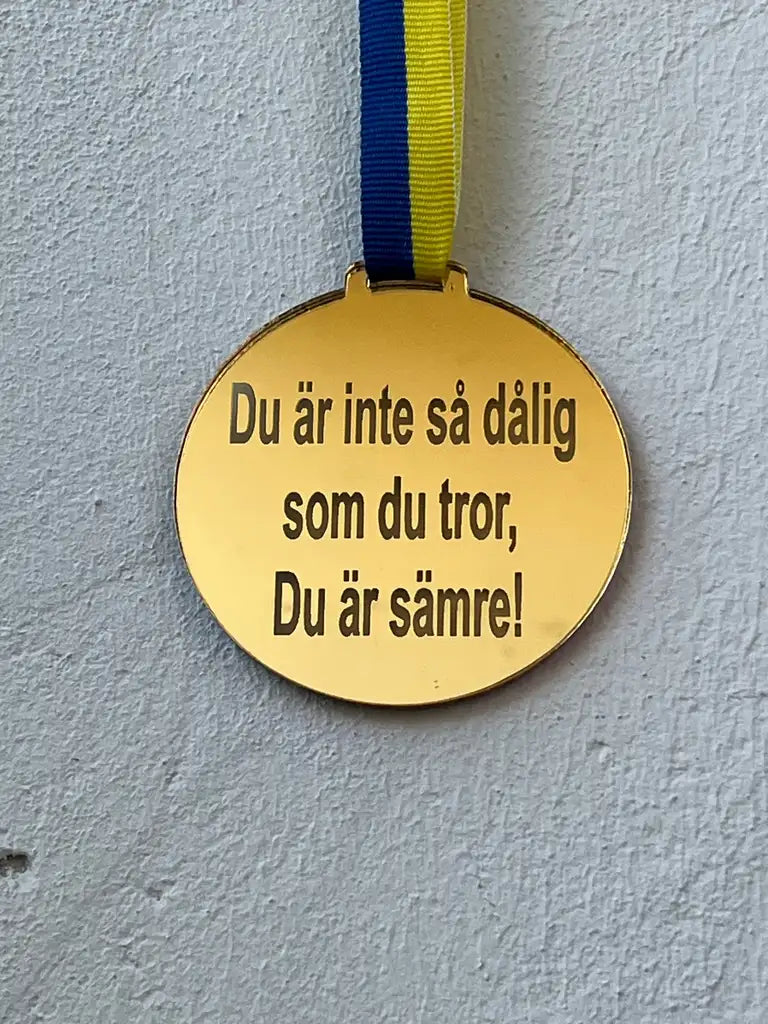 Medalj | Du är inte så dålig som du tror, Du är sämre! från Express-presenter har en spegelplast guld finish, blågult band och är graverad med svensk text mot en grå vägg.