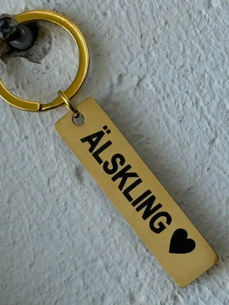 Nyckelringen | Älskling by Express-presentatör är en nyckelring i guld med en rektangulär tagg, med laseretsat "ÄLSKLING" och ett svart hjärta, som visas hängande mot en vit vägg med textur.