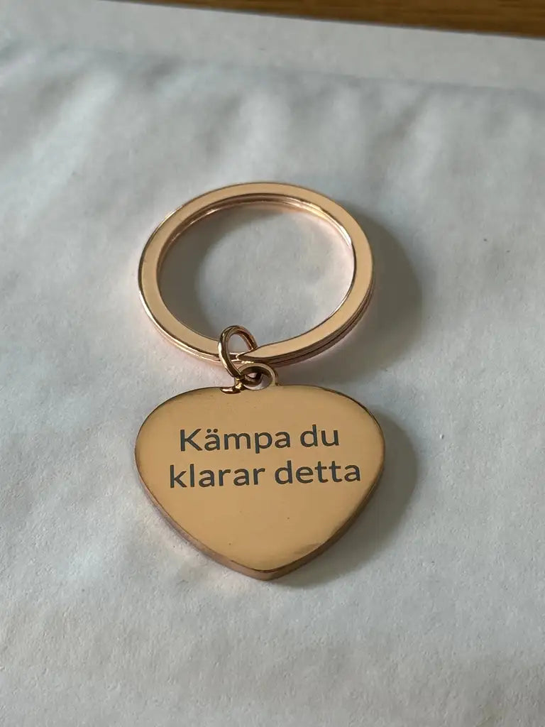 Nyckelringen | Kämpa du klarar detta från Express-presenter är en guldfärgad, hjärtformad nyckelring med lasermärkt text "Kämpa du klarar detta" på vit yta.