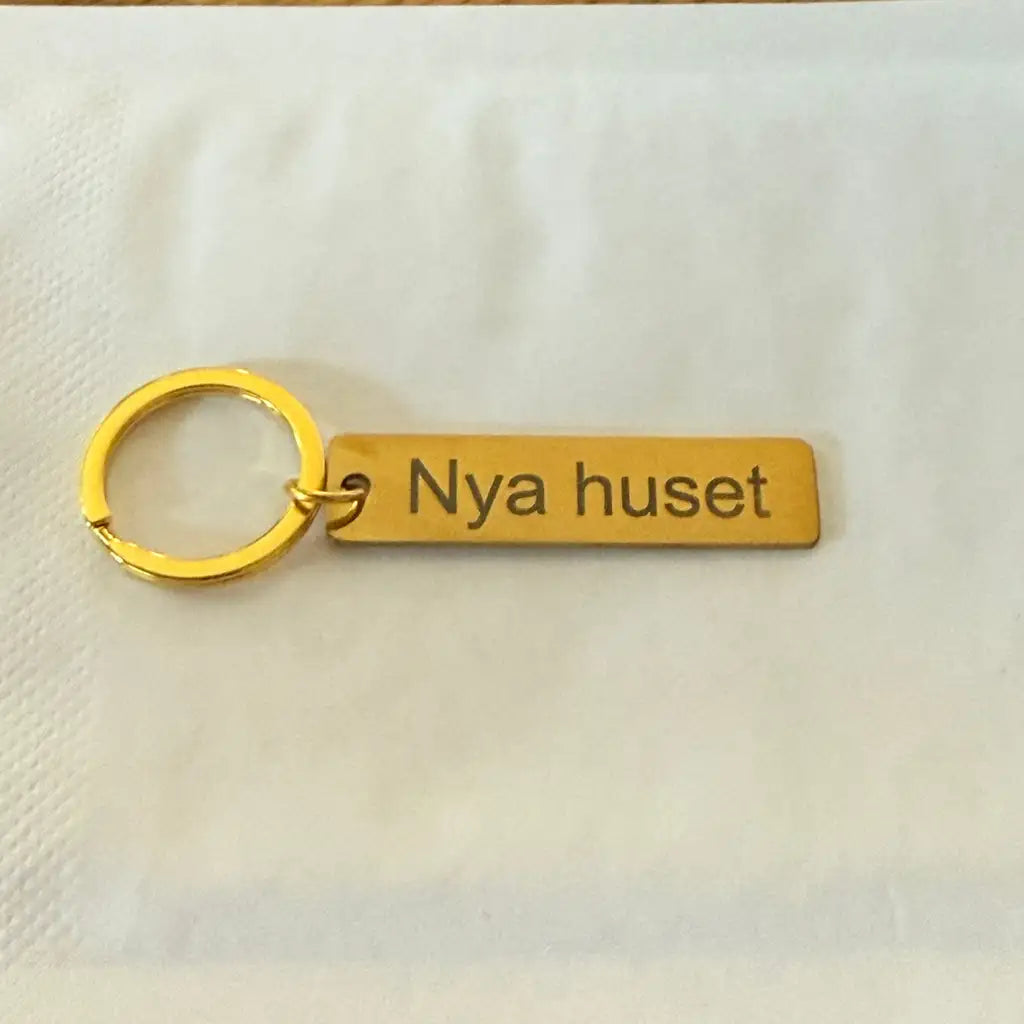 Express-presenter's Nyckelring | Nya huset är en nyckelring i guld med en rektangulär tagg graverad med "Nya huset" på en vit yta - en perfekt och elegant inflyttningspresent.