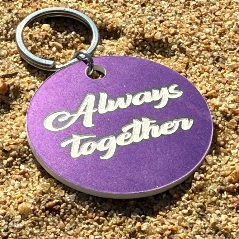 En rund, mörklila 40mm Nyckelring | Always together by Express-presenter med "Always together" ingraverat i vit kursiv text ligger på sandig mark.