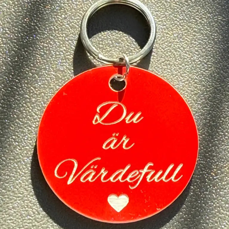 Nyckelringen Du är värdefull (röd) från Express-presenter är en 40 mm röd nyckelring med "Du är Värdefull" och ett vitt hjärta, på en grå yta med struktur.
