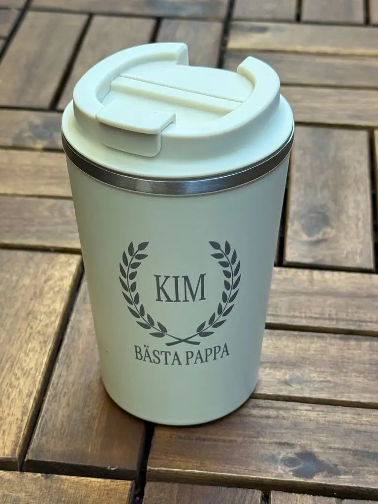 På ett träbord står en vit Termosmugg | Bästa pappa från Express-presenter. Den är tillverkad av rostfritt stål, har "KIM" och "BÄSTA PAPPA" lasergraverade med en lagerkransdesign och kommer med ett lock.