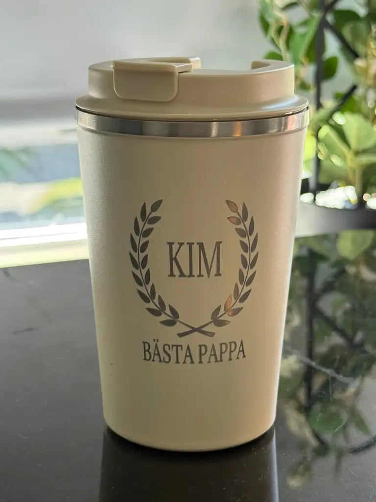 Termosmugg | Bästa pappa från Express-presenter, en resemugg i beige rostfritt stål med "Kim" och "Bästa Pappa" i lasergravyr omgivet av en lagerkrans, sitter på en reflekterande yta med gröna växter i bakgrunden.