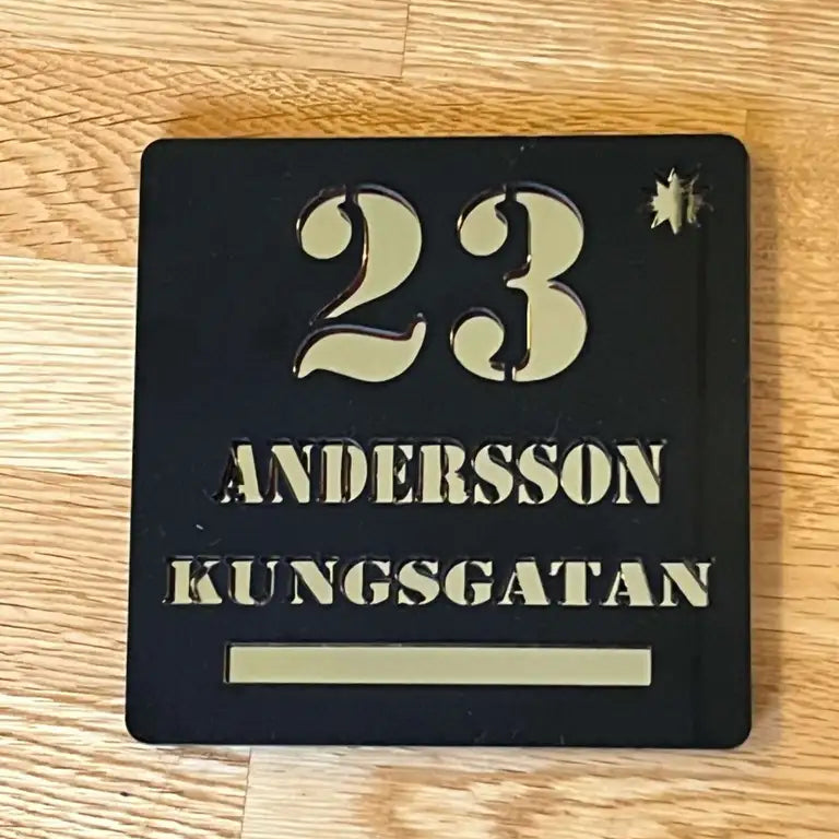 Skylten Skyltar för ditt hus av Express-presenter, med siffran 23 högst upp och namnen ANDERSSON och KUNGSGATAN nedanför i svart och guld 6mm akryl, är placerad på ett trägolv.