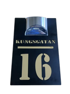 En svart rektangulär Husskylt solcellsdriven från Express-presenter med "KUNGSGATAN 16" i guld och en silverfärgad LED-solcellslampa på toppen för dekorativ belysning.