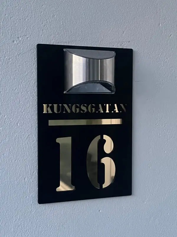En svart och guldfärgad Husskylt solcellsdriven av Express-presenter är monterad på en ljusgrå vägg och visar "KUNGSGATAN 16" med en silverfärgad dekorationslampa ovanför för ett modernt utseende.