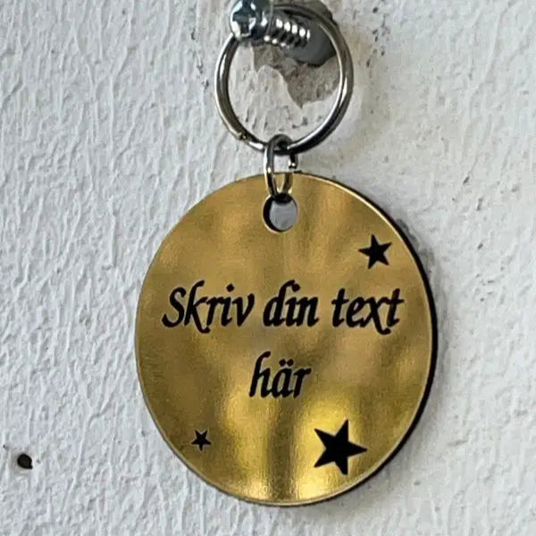 Nyckelringen med egen text från Expresspresenter.se är en 40 mm guldfärgad nyckelring med svensk text "Skriv din personliga text här" och små svarta stjärnor, som visas hängande på en vägg.