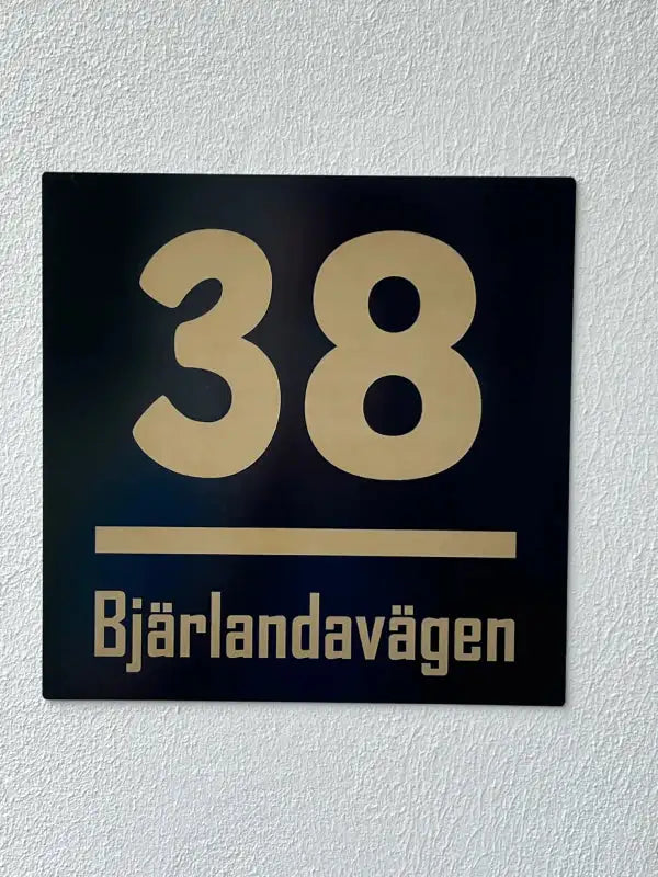 Express-presenter Husskylt nummer har guldtext med husnummer 38 över en rad, "Bjärlandavägen" under och är monterad på en vit texturerad vägg.