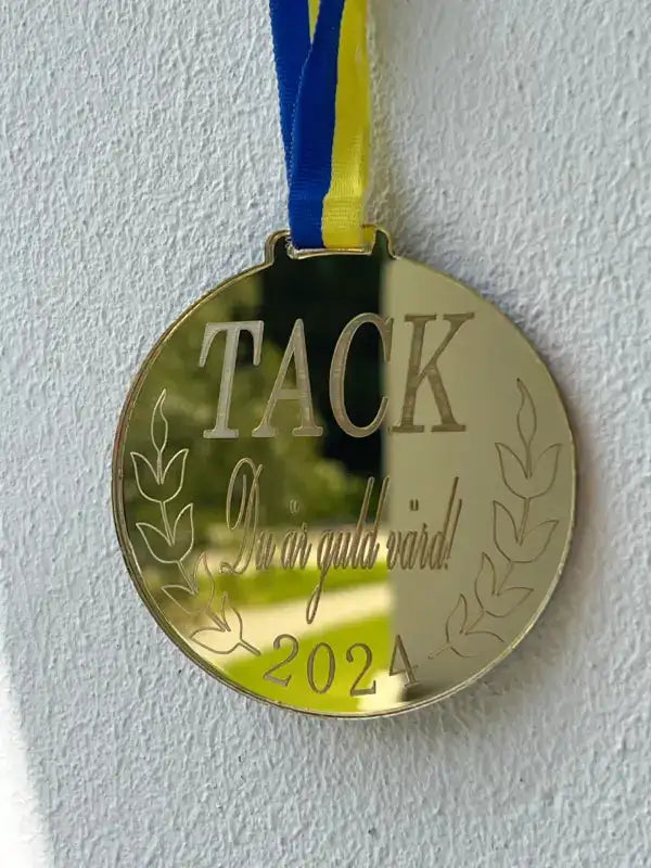 Medalj Du är guld värd från Express-presenter har en spegelplast guldfinish graverad med "TACK Du är guld värd! 2024" och lagerkvistar, hängande i ett blågult band mot en vit vägg.