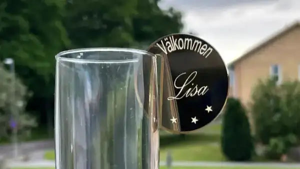 Express-presenter Dekorera festen med glasmarkörer har en rund spegel akryl tagg graverad med "Välkommen Lisa" och tre stjärnor, fäst på ett glas - perfekt som personlig etiketter eller glasmarkörer för ditt evenemang. Oskarp grönska och byggnader i bakgrunden.