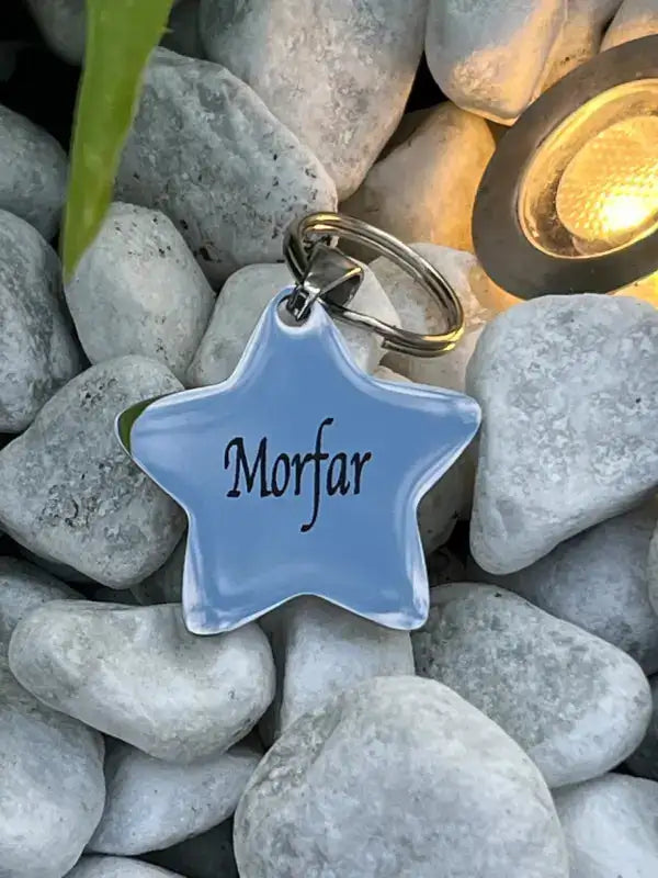 Nyckelringen med texten Morfar från Expresspresenter.se, formad som en stjärna, vilar på vita stenar vid en glödande trädgårdslampa - en perfekt present.