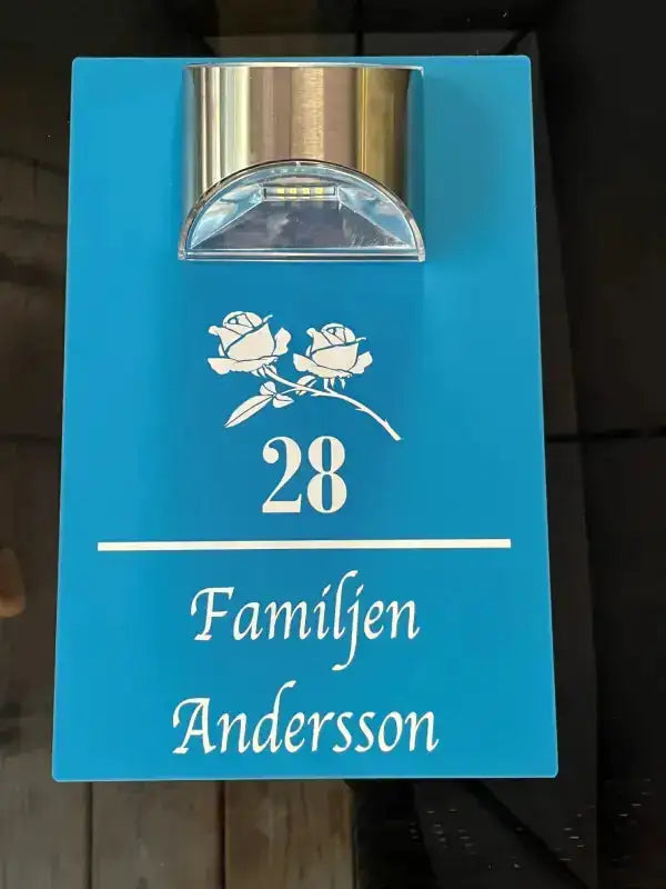 Express-presenter Hus nummerskylt med namn och belysning: En blå skylt med silvrig taklampa, två vita rosor, siffran 28 samt texten "Familjen Andersson" i vitt.