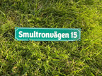 En Adresskylt från Express-presenter, med grön bakgrund och vit text "Smultronvägen 15", mäter 300×100 mm med skruvhål och visas liggande på ljusgrönt gräs.
