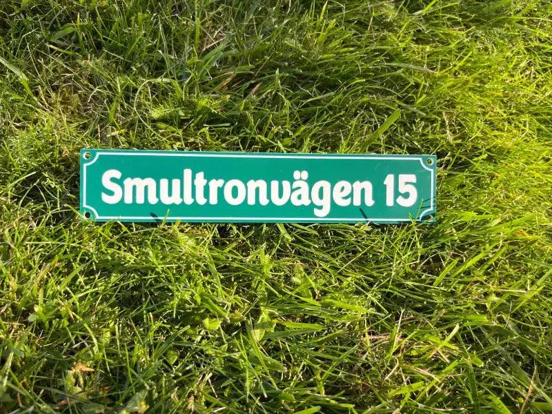 En Adresskylt från Express-presenter, med grön bakgrund och vit text "Smultronvägen 15", mäter 300×100 mm med skruvhål och visas liggande på ljusgrönt gräs.