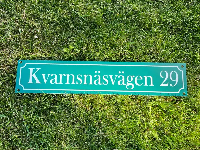 En Adresskylt från Express-presenter, rektangulär och grön med vit text "Kvarnsnäsvägen 29", 300×100 mm, ligger i gräset.