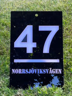 Fasadskylt exklusiv från Express-presenter är en svart akrylskylt med "47" och "NORRSJÖVIKSVÄGEN" i vitt, som står upprätt på grönt gräs som en elegant husnummerskylt.