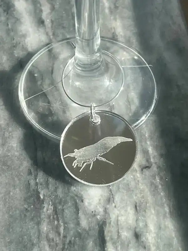 Dekoration glasmarkör till räkafton från Expresspresenter.se - en rund vinglasmarkör i silver med räkdesign - hänger elegant från skaftet, perfekt för din nästa räkafton och visas på en gråmarmorerad yta.