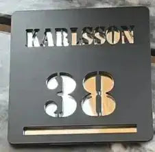 Brevlådeskylt by Express-presenter är en svart brevlådeskylt med "KARLSSON" utskuret upptill, siffran 38 nedtill och en smal horisontell springa nedtill.