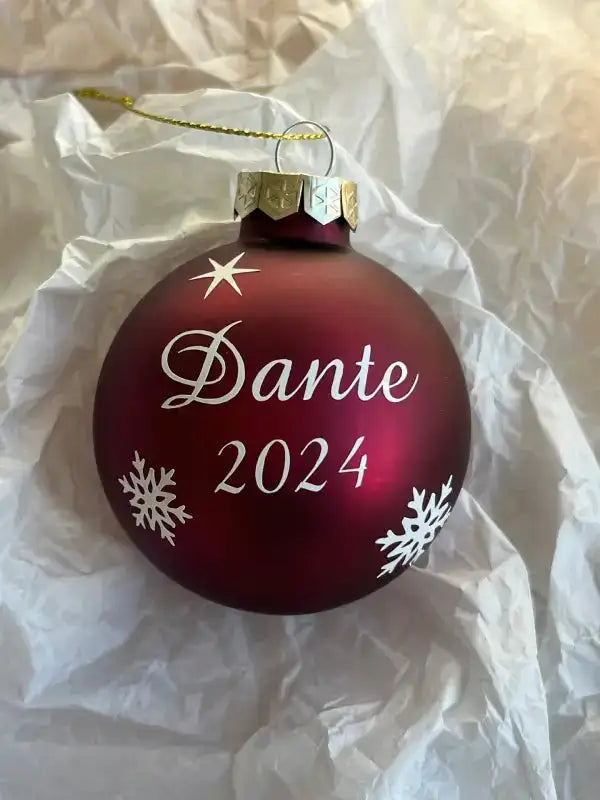 Den mörkröda julkulan i glas från Expresspresenter.se, personligt dekorerad med namnet Dante och året 2024, har vita snöflingor och en stjärna och vilar på vitt silkespapper.