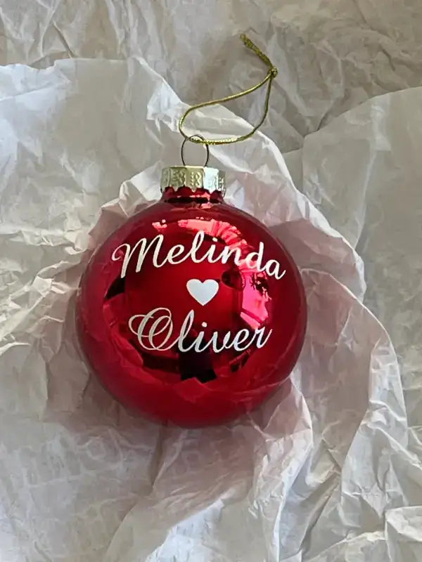 A glänsande röd Personlig julpresent med text från Expresspresenter.se med "Melinda ♥ Oliver" i vit skrift vilar på vitt silkespapper-en perfekt och charmig julgranskula med namn till din gran.