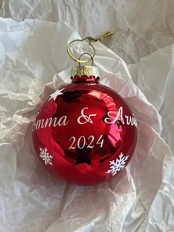 A Personlig julgranskula med text från Expresspresenter.se - röd, glansig och dekorerad med vita snöflingor samt texten Emma & Arun 2024 i skrivstil, placerad på skrynkligt vitt silkespapper.