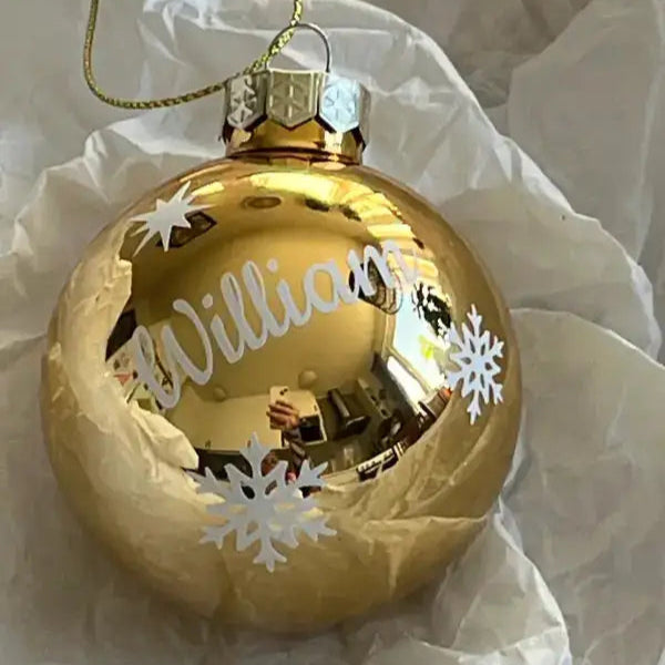 En glänsande Julgranskula i tomt guld med egen text från Expresspresenter.se, dekorerad med vita snöflingor och namnet William, vilar på skrynkligt vitt silkespapper.