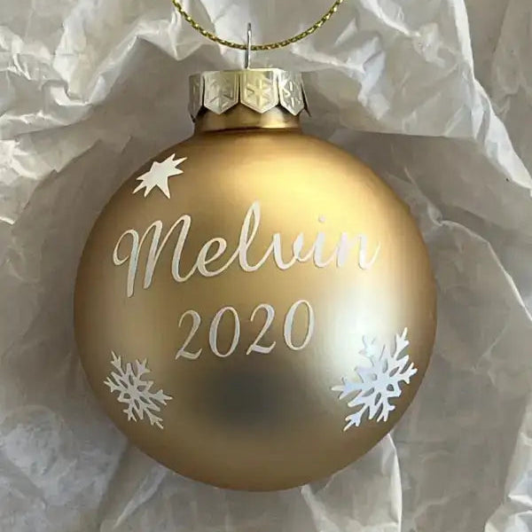 A Julgranskula i guld med egen text från Expresspresenter.se (storlek 6 cm), här med texten "Melvin 2020" och vita snöflingor, vilar på skrynklat vitt silkespapper.