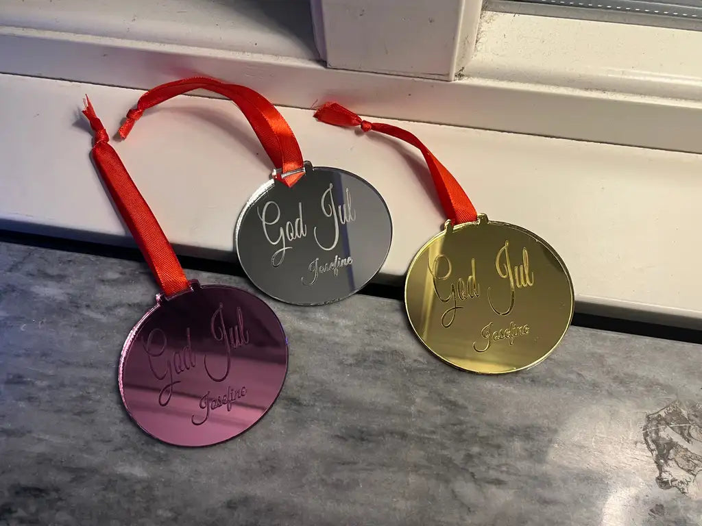 Tre runda Julgranshänge | God Jul by Express-presenter - ett rosa, ett silver och ett guld - med blanka ytor, graveringen "God Jul Josefine" och röda band, står på en fönsterbräda.