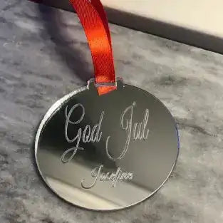 Express-presenter's Julgranshänge | God Jul är en rund julgranskula i silver med "God Jul" och Josefine ingraverat, upphängd i ett rött band och placerad på en grå marmoryta.