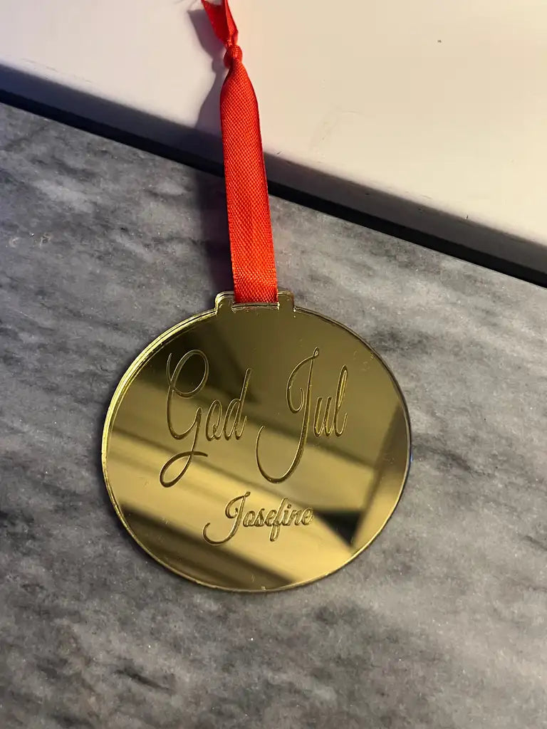 Express-presentatören Julgranshänge | God Jul, en oval guldprydnad med röd rosett och graverat "God Jul" samt namnet Josefine, är placerad på en grå marmoryta med en vit kant.