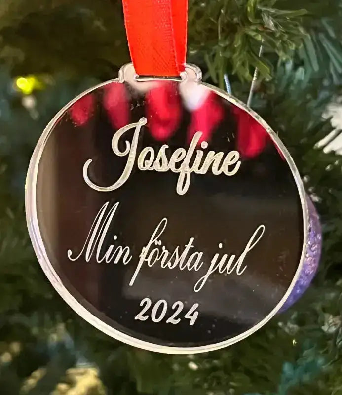Ett runt, reflekterande Julgranshänge | Min första jul by Express-presenter, 70 mm stort och graverat med "Josefine Min första jul 2024", hänger i en gran med ett rött band.